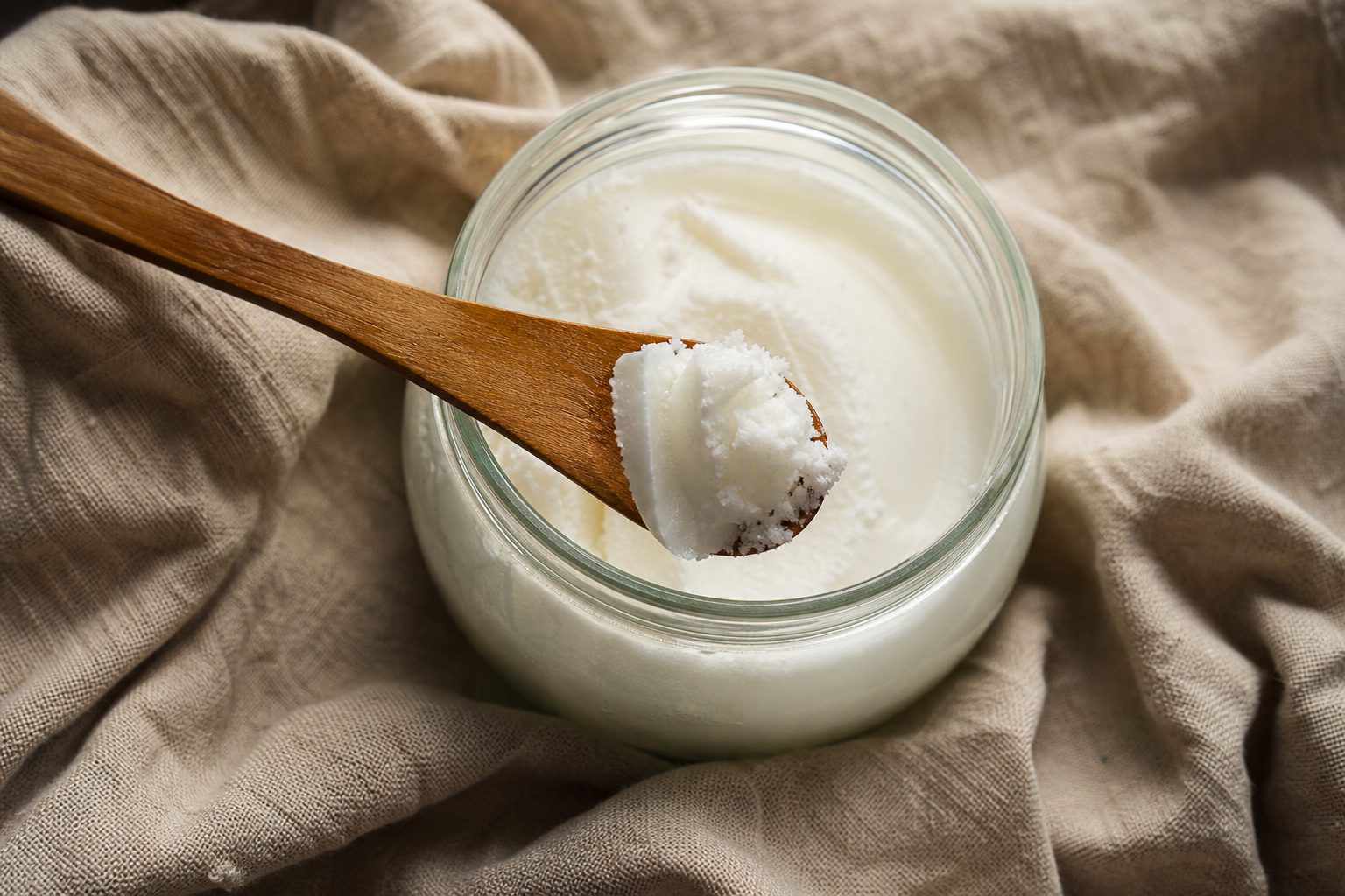 An open glass jar of solid white coconut oil on a linen cloth, with a wooden spoon over the jar holding a scoop of the oil